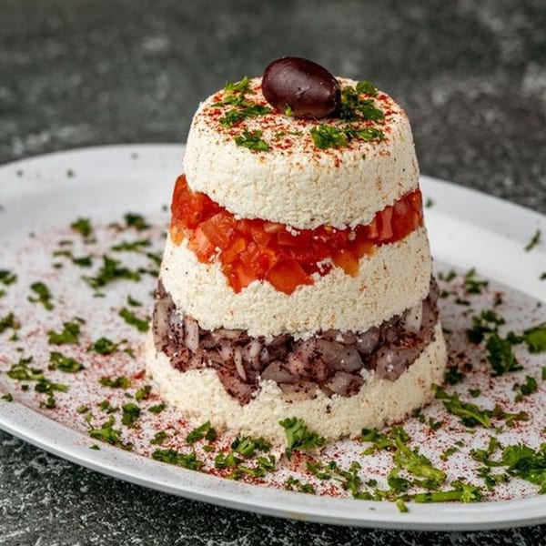 Layered Mediterranean dip with white cheese, red tomatoes, dark olives, and herbs on a white plate.