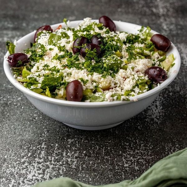 A white bowl of Mediterranean salad with lettuce, feta cheese, parsley, and whole Kalamata olives.
