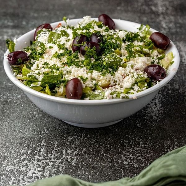 A white bowl filled with a Mediterranean salad, topped with feta cheese, parsley, and olives.