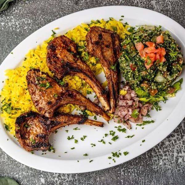 Four grilled lamb chops with yellow rice, tabbouleh salad, and diced onions on a white platter.