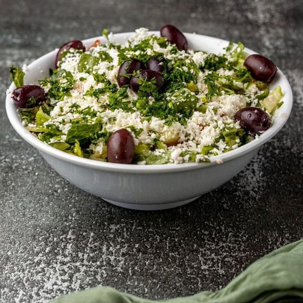 A white bowl filled with a Mediterranean salad, topped with feta cheese, parsley, and Kalamata olives.