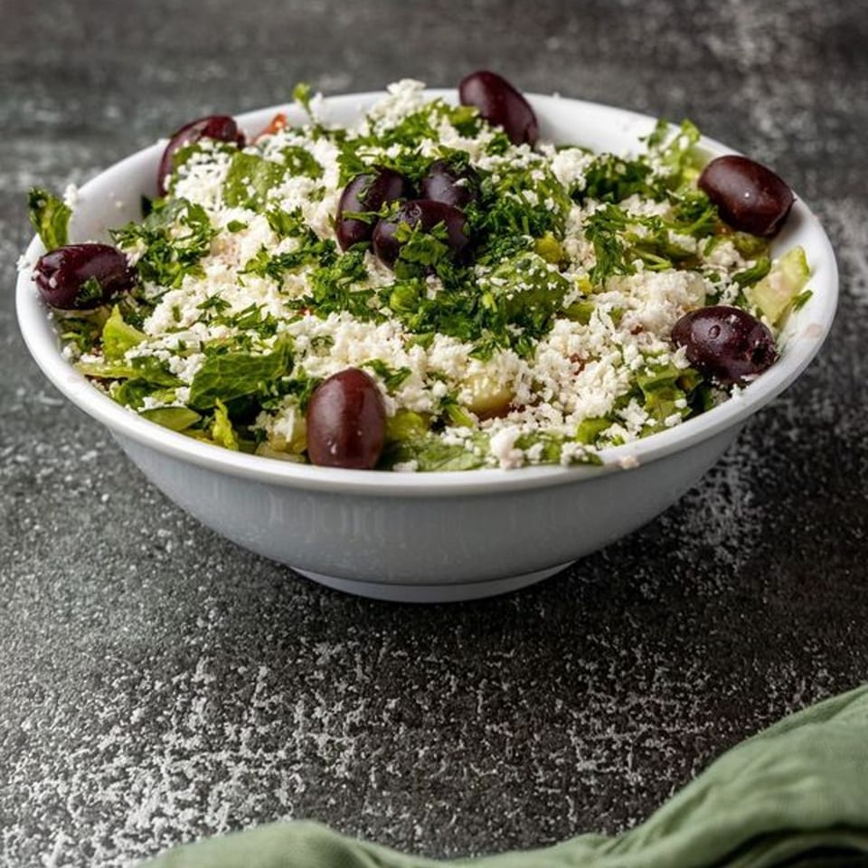 A white bowl filled with a Mediterranean salad, topped with feta cheese, parsley, and Kalamata olives.