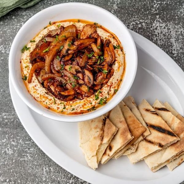A white bowl of hummus topped with sautéed mushrooms and onions, served with grilled pita bread.