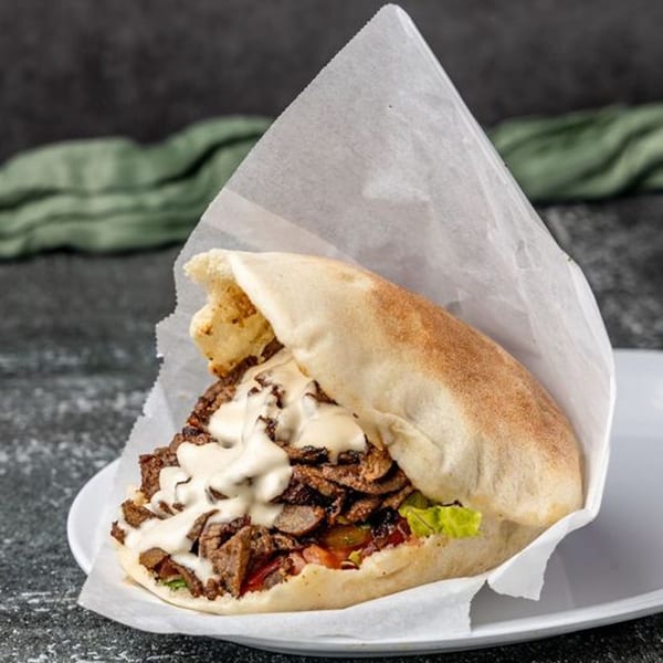 A Pita Shop pita sandwich filled with meat, lettuce, tomatoes, and white sauce, wrapped in paper.