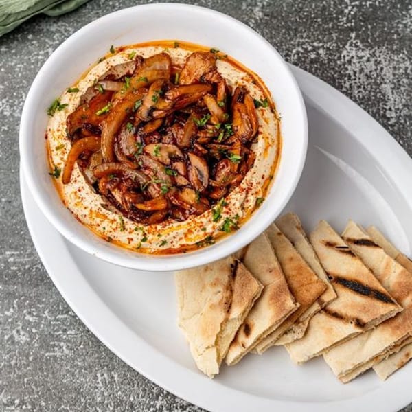 A white bowl of Pita Shop hummus topped with sautéed mushrooms and onions, served with pita bread.