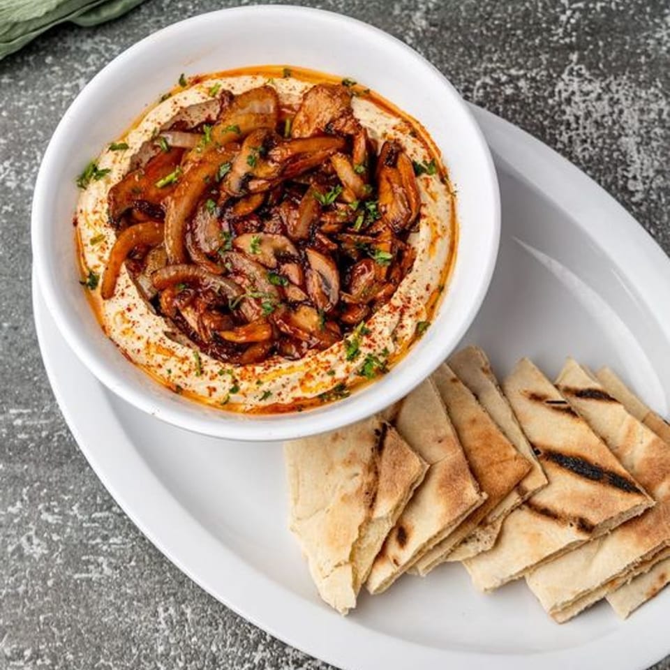 A white bowl of Pita Shop hummus topped with sautéed mushrooms and onions, served with pita bread.