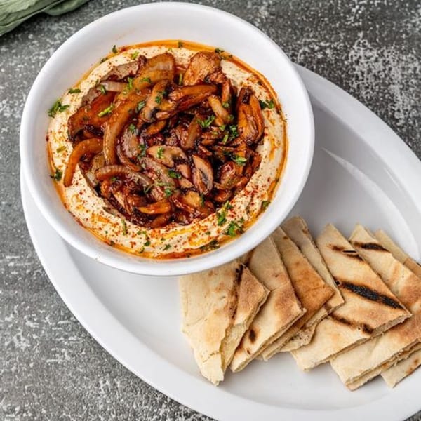 A white bowl of hummus topped with sautéed mushrooms and onions, served with pita bread.